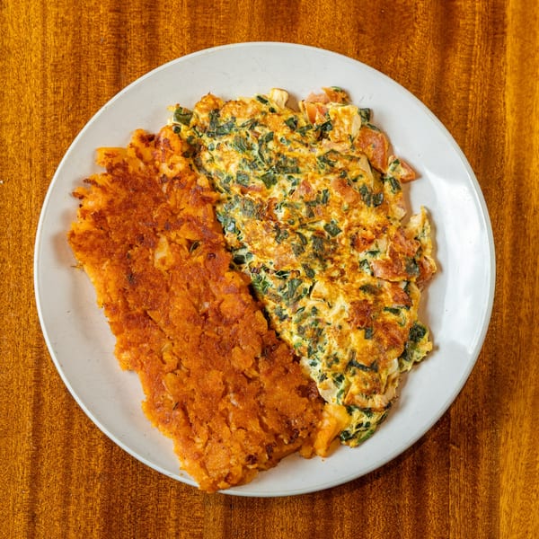A white plate holds a golden-brown hash brown and a fluffy omelet with spinach and tomatoes.
