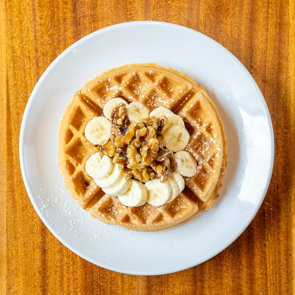 Golden waffle topped with sliced bananas, candied walnuts, powdered sugar, and syrup on a white plate.